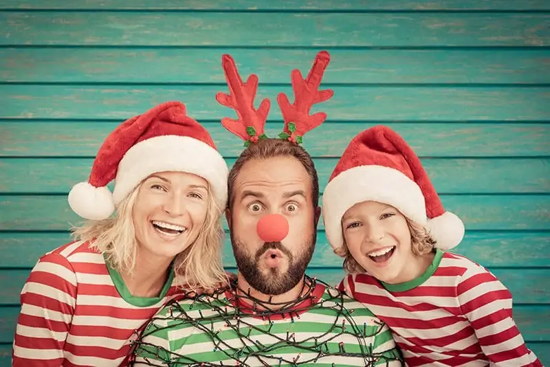 Two adults and a child in festive striped shirts, Santa hats, and reindeer antlers smiling in front of a wall.